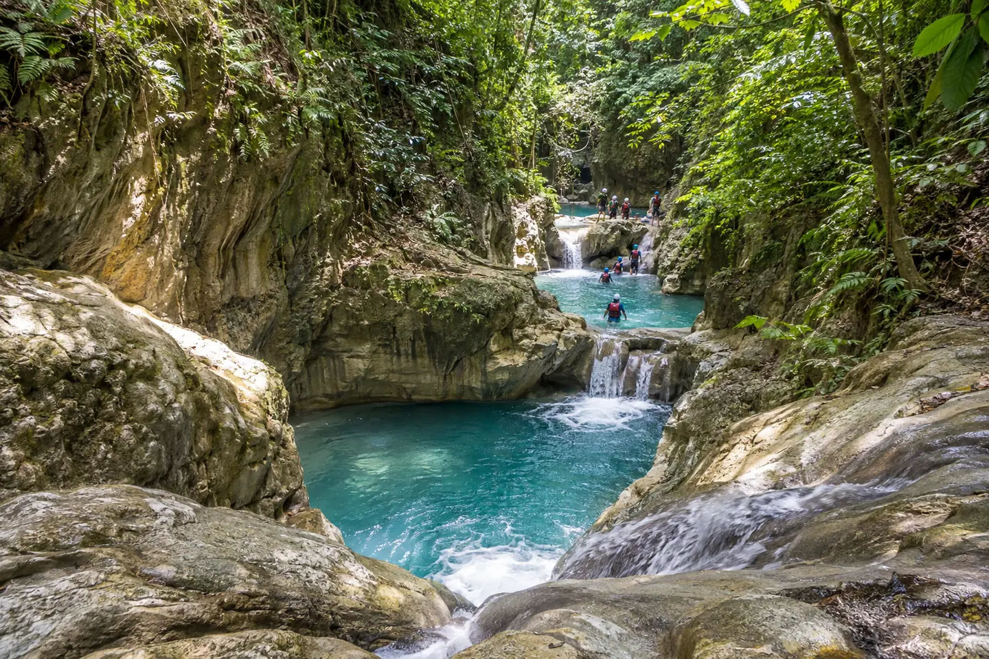 Guided visitors in helmets and life vests beside a clear waterfall pool at Damajagua